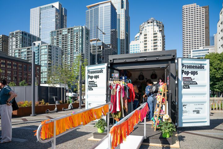 A shipping container converted into a Promenade Pop-Up shop with colorful clothing displays and the Seattle skyline behind it.