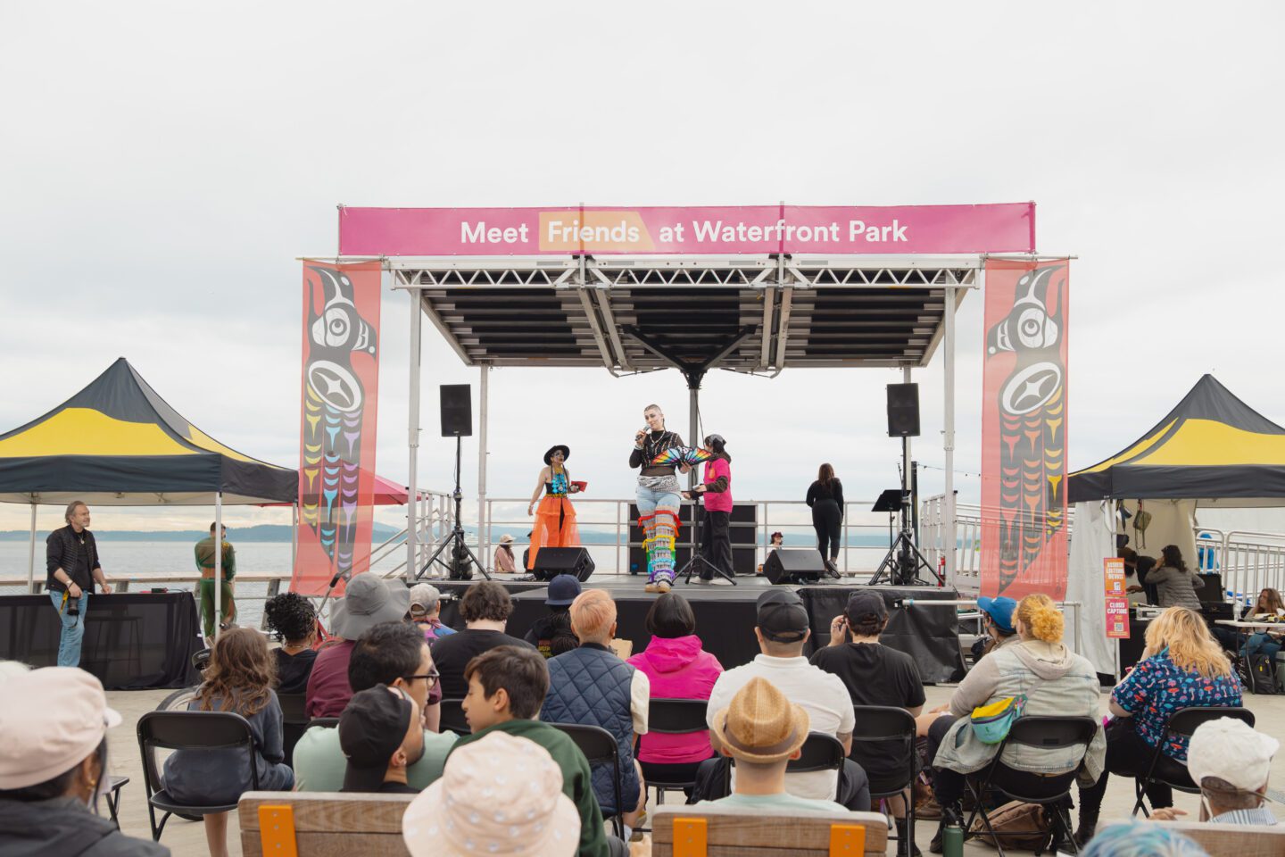 Three individuals on stage, speaking towards a crowd. They are on a stage with banners on the top and sides that read Meet Me at Waterfront Park and Indigenous design. There are two black and yellow tents on each side of the stage