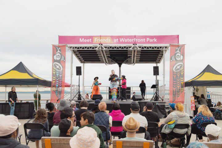 Three individuals on stage, speaking towards a crowd. They are on a stage with banners on the top and sides that read Meet Me at Waterfront Park and Indigenous design. There are two black and yellow tents on each side of the stage