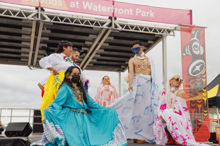 A group of six fashion show models are on stage facing towards a crowd.