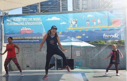 A woman in a black tank top and cap jumps mid-air on an outdoor stage, flanked by two other dancers, with a bright Friends of Waterfront Park banner and the city skyline behind them