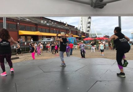 Zumba instructors lead a class from an outdoor stage while a crowd of participants dance on the wooden pier boardwalk, with the Seattle Great Wheel and Elliott Bay visible in the distance.]