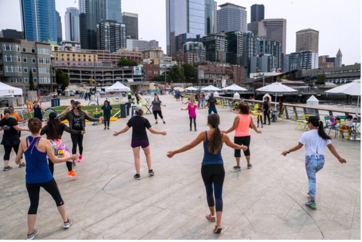 A group of people spread out in a circle on an open plaza at Pier 62, arms extended mid-dance, with white umbrellas, yellow chairs, and the Seattle skyline visible behind them on an overcast day.]