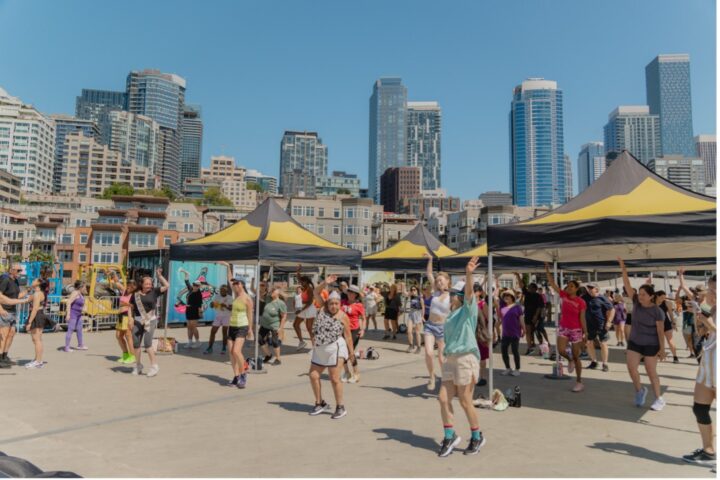A large group of people dance together on a sunny plaza at Waterfront Park, arms raised, with the Seattle skyline rising behind them.
