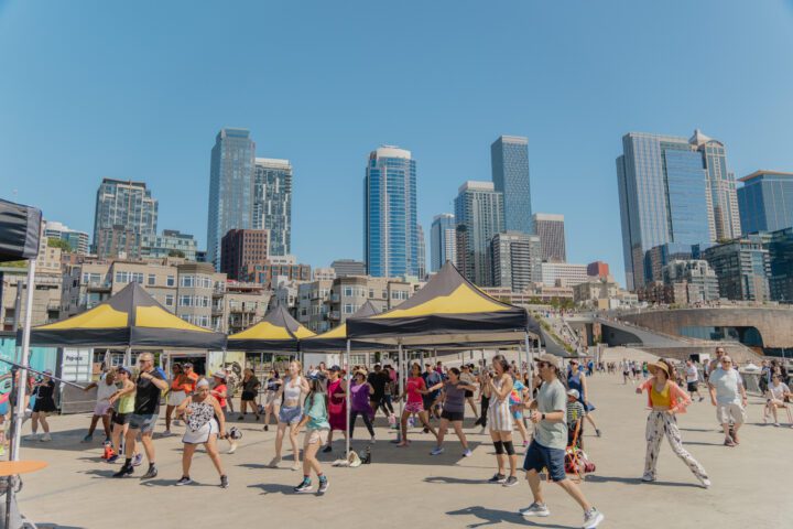 A large group of smiling participants stretch their arms wide mid-dance on Pier 62, with the Seattle skyline and Great Wheel visible in the background on a bright summer day.