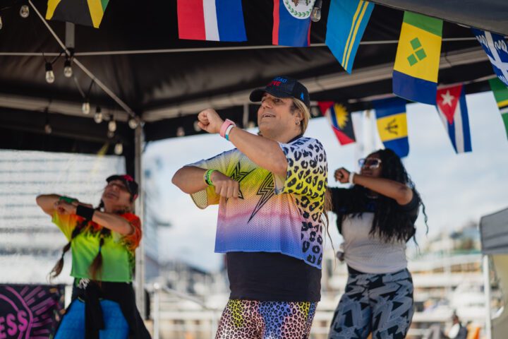 A Zumba instructor in a bright yellow and white outfit dances on an outdoor stage with arms outstretched, surrounded by colorful international flags hanging from the tent above on a sunny day.