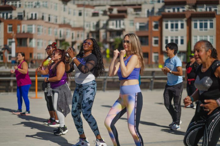 A diverse group of smiling participants dance together on an open waterfront plaza in colorful workout gear, with residential buildings behind them.