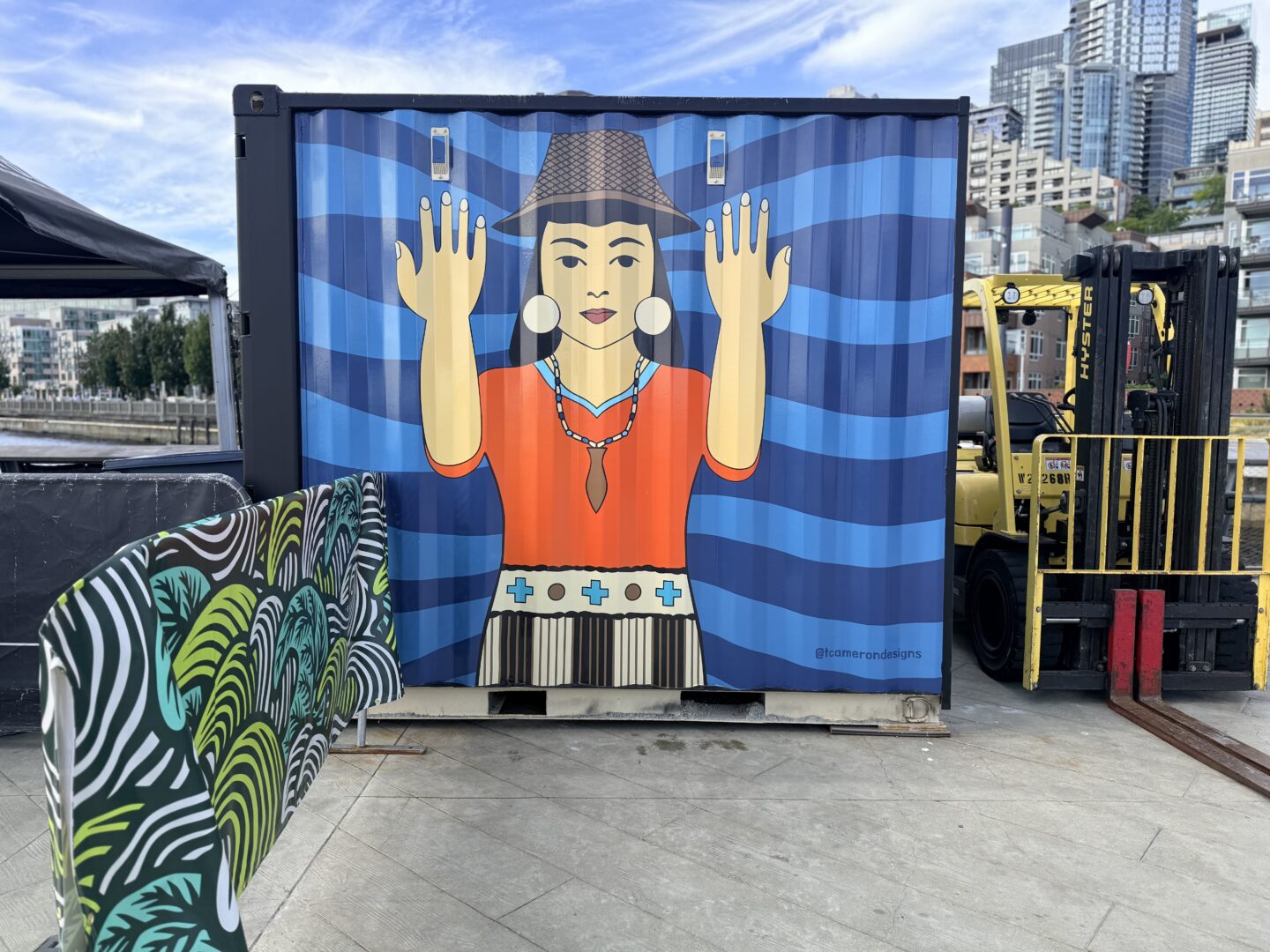 A shipping container on Pier 62 featuring a welcoming figure in traditional regalia with raised hands, set against a blue wave background — part of Taylor Cameron's Currents of Connection mural during installation.