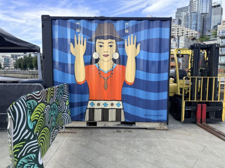 A shipping container on Pier 62 featuring a welcoming figure in traditional regalia with raised hands, set against a blue wave background — part of Taylor Cameron's Currents of Connection mural during installation.