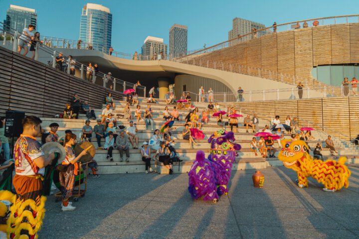 Crowd of people sit on the cedar steps, which are form in amphitheater style. Performers are on the ground level with two of them in dragon attire. It’s golden hour with the sun setting.