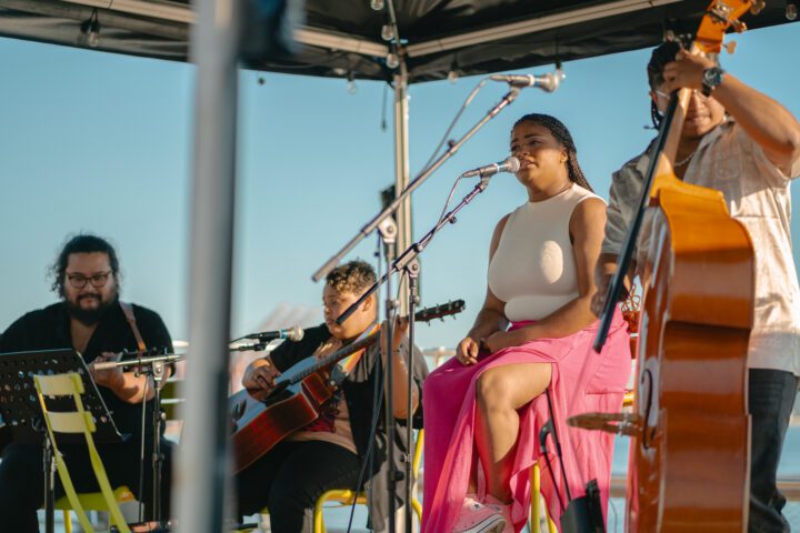 Four performers on stage with three sitting on stools, person in the middle singing into microphone and person on the right playing the cello.