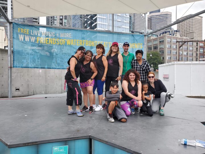 A group of about ten people in Zumba attire pose together on an outdoor stage on Pier 58, smiling for the camera with the Seattle skyline behind them and a Friends of Waterfront Seattle banner in the background