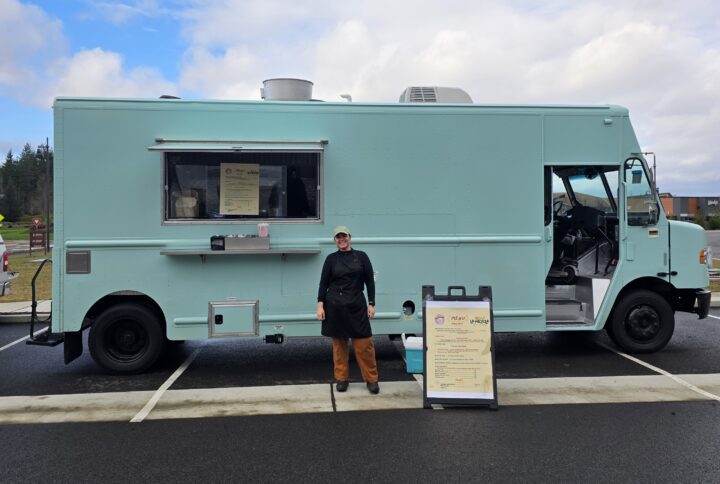 A woman in a black chef's jacket and apron stands in front of a large mint green food truck with a menu board displayed beside her, smiling at the camera under a partly cloudy sky.