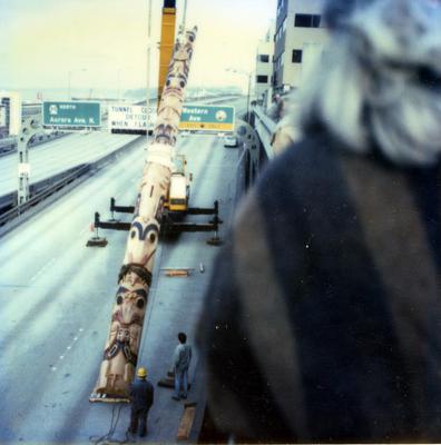 A person from the back watching a large totem pole being transported on a highway.