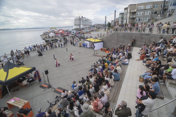 A large crowd sitting at the Salish Steps watching two performers with Pier 62 in the background.