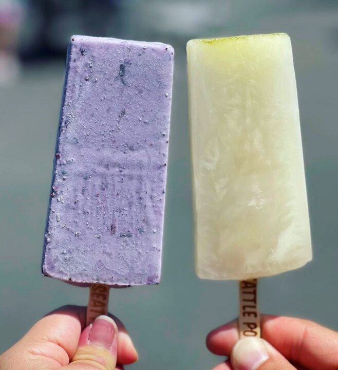 Two hands holding up popsicles, one purple and one light yellow.