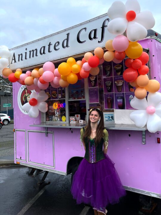 A smiling woman in a purple tulle skirt and green corset top stands in front of a purple food truck decorated with a colorful balloon arch, with the Animated Cafe sign visible behind her.