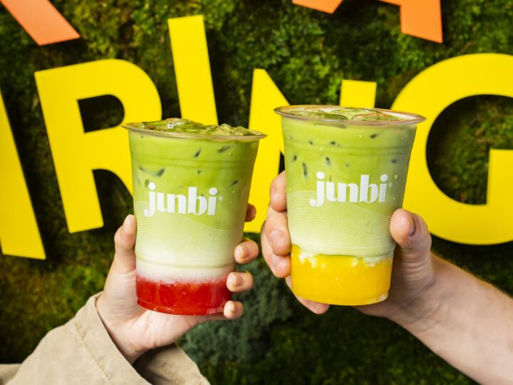 wo hands hold up vibrant layered iced matcha drinks in Junbi-branded cups, with bold yellow letters and a green backdrop behind them. 