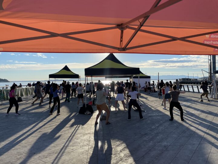 A group of people facing the water dancing along at an outdoor zumba class.
