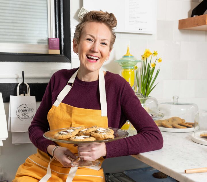 A person wearing an orange apron smiling holding a tray of cookies.