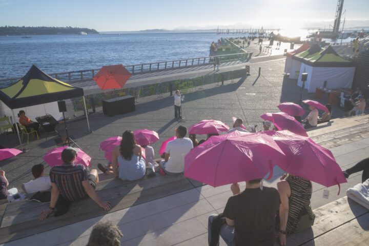 A group of people sitting on the Salish Steps holding pink umbrellas watching a performer.