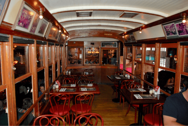 Inside of a tram car with windows on either side and red tables and chairs throughout.