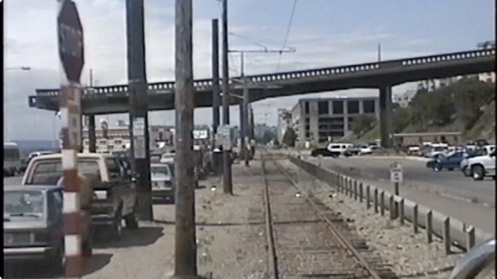 A photo looking down a set of train tracks with cars parallel parked on the left and a large parking lot on the right.